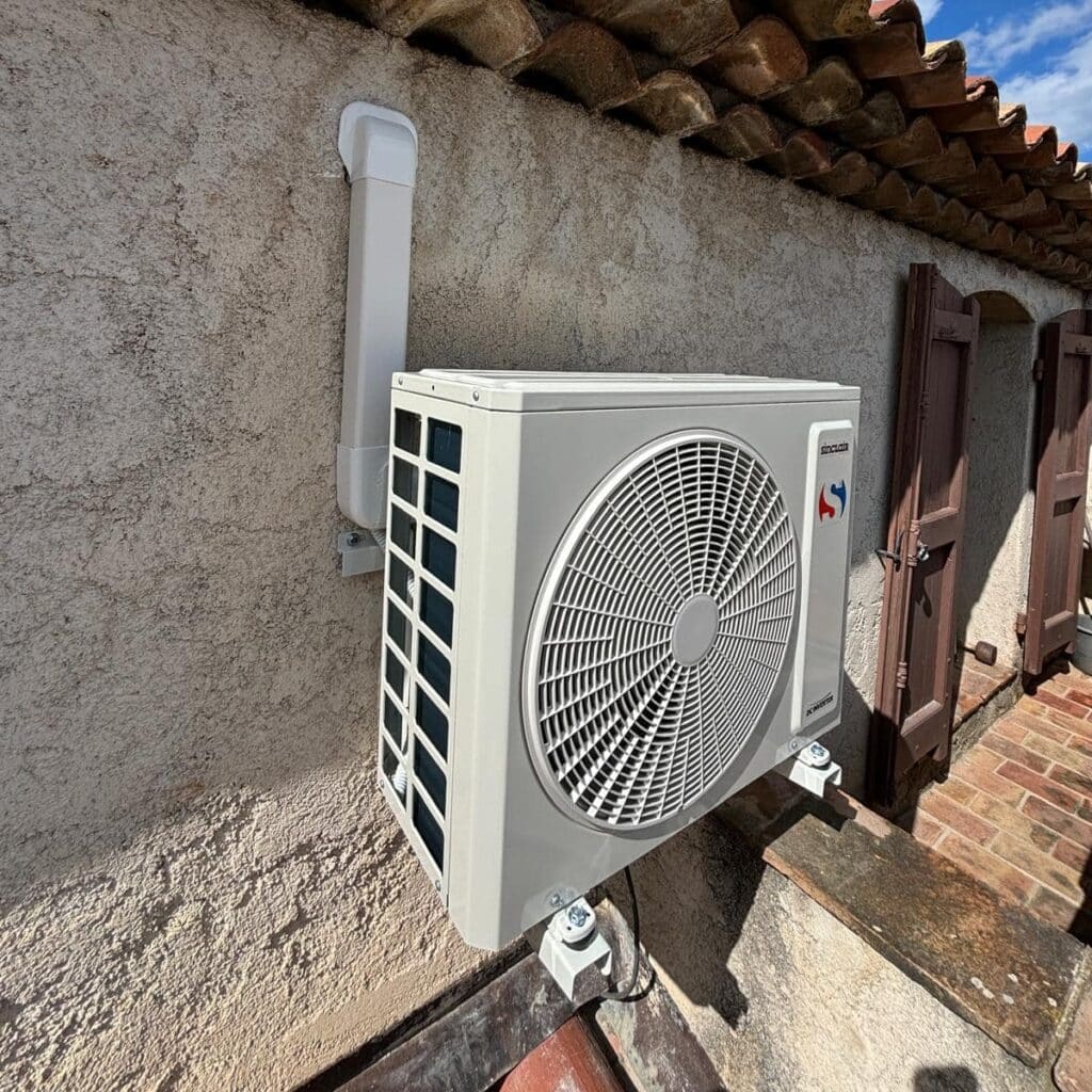 Climatiseur Sinclor DC Inverter blanc installé sur mur crépi avec goulotte, sous des tuiles de toit et à côté de volets en bois.