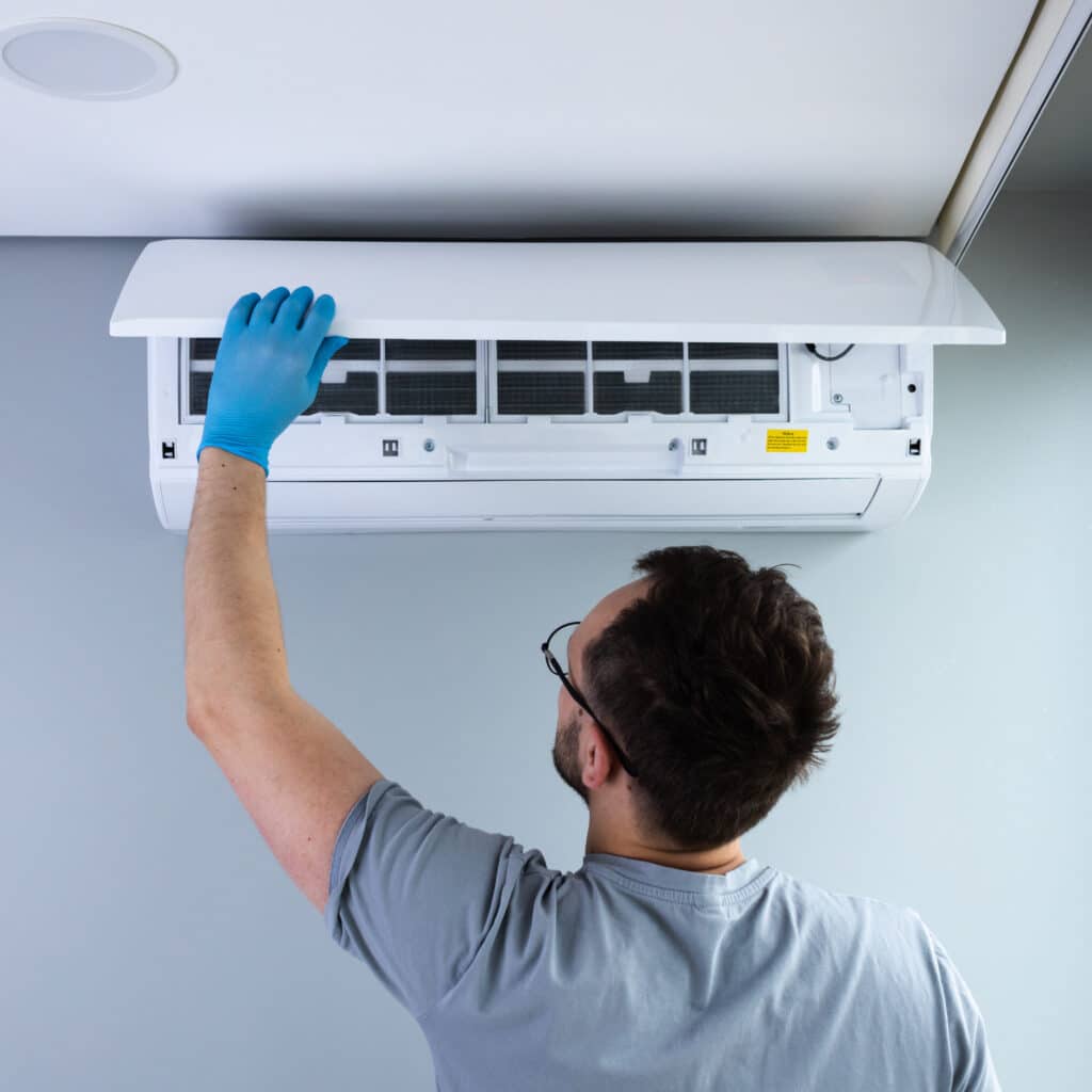 Homme ganté inspectant un climatiseur mural ouvert, filtres visibles. Autocollant jaune « Notice » à l'intérieur. Entretien de la climatisation.