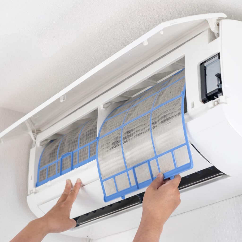 Mains retirant un filtre à air encrassé (cadre bleu) d'un climatiseur mural blanc. Entretien essentiel pour un air sain.