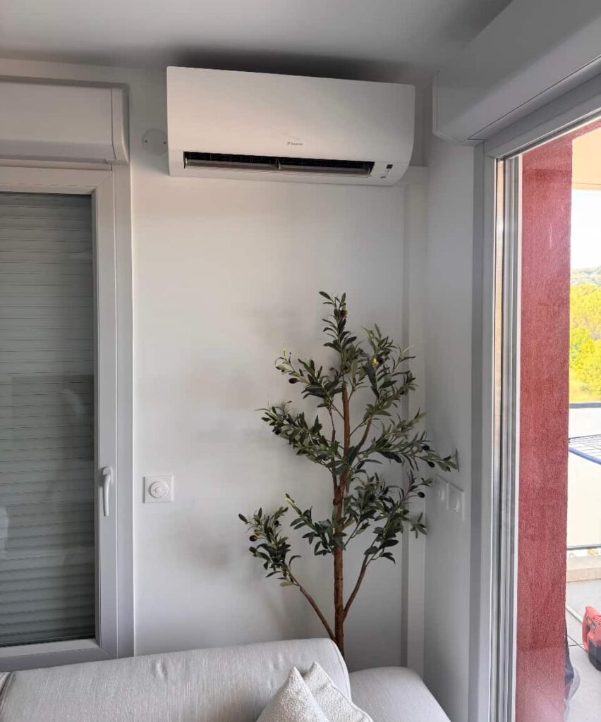 Climatiseur Daikin mural blanc dans un salon moderne avec une plante olivier artificielle près d'une fenêtre et un canapé clair.