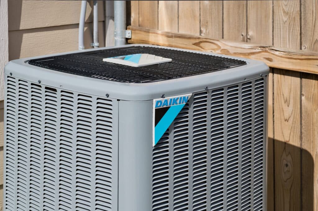 Unité extérieure de climatiseur Daikin grise, avec grille supérieure noire et logo Daikin bleu et noir, contre une clôture en bois.
