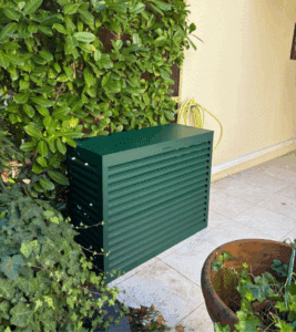 Cache-groupe extérieur de climatisation vert foncé avec lames horizontales, dissimulé près d'un mur jaune pâle et d'un feuillage luxuriant, avec un pot en terre cuite.