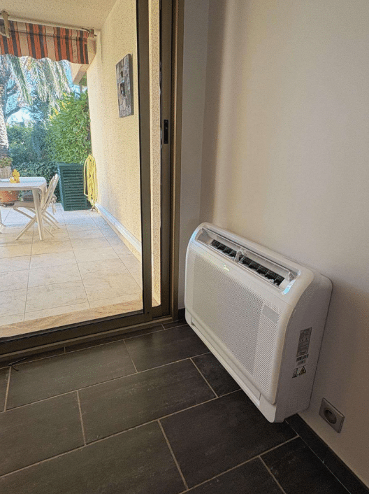 Climatiseur Midea gainable au sol, près d'une porte-fenêtre coulissante donnant sur une terrasse ensoleillée avec table et végétation.